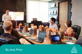 A man standing in front of a screen presenting to a table full of people with laptops in front of them