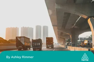 Sunset view of a busy highway under an elevated road, with trucks driving and high-rise buildings in the background.