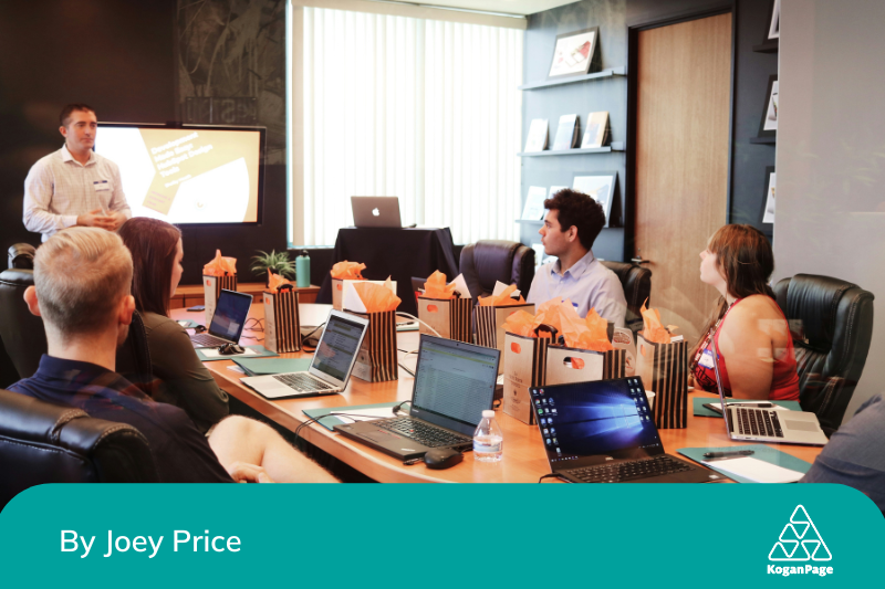 A man standing in front of a screen presenting to a table full of people with laptops in front of them