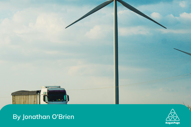 A large wind turbine stands in a field under a partly cloudy sky. A truck is driving along a road in front of the wind turbine.