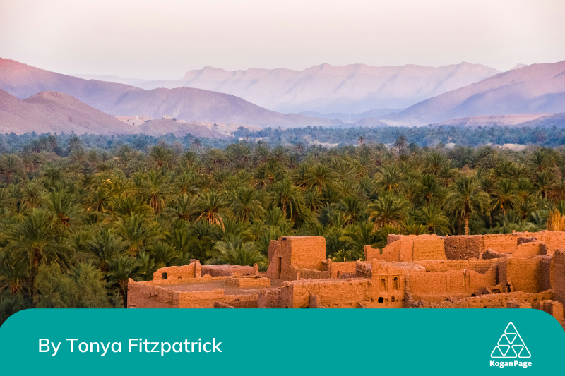 A picture of clay buildings next to an oasis with a Moroccan mountain range in the background 